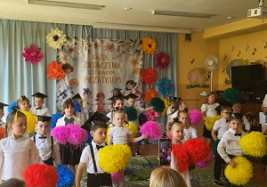 Przedszkolaki podczas występu tanecznego z kolorowymi pomponami na uroczystości zakończenia przedszkola. W tle widoczna jest barwna dekoracja i okolicznościowy napis.