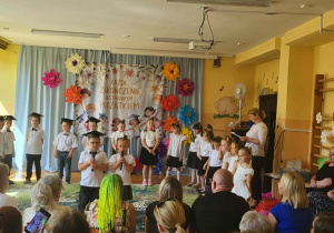 Dzieci z grupy starszaków prezentują program artystyczny podczas uroczystości zakończenia przedszkola. Przedszkolaki stoją na tle dekoracji z kolorowymi kwiatami i banerem z napisem „Każde zakończenie jest nowym początkiem”. Dwoje dzieci na środku trzyma mikrofony i występuje przed zgromadzonymi rodzicami oraz gośćmi. Chłopcy ubrani są w białe koszule i papierowe birety, a dziewczynki w białe bluzki i ciemne spódniczki. W dolnej części zdjęcia widoczni są uczestnicy uroczystości obserwujący występ.