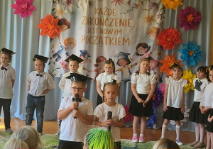Przedszkolaki podczas występu artystycznego z okazji zakończenia przedszkola. Dzieci prezentują program słowno-muzyczny na tle kolorowej dekoracji i okolicznościowego napisu.