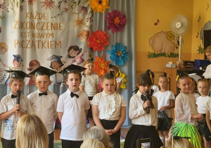 Przedszkolaki podczas występu artystycznego z okazji zakończenia przedszkola. Dzieci prezentują program słowno-muzyczny na tle kolorowej dekoracji i okolicznościowego napisu.