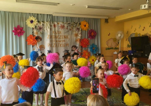 Dzieci wykonują choreografię z kolorowymi pomponami podczas programu artystycznego przygotowanego na zakończenie roku przedszkolnego. Występ odbywa się przed rodzicami i zaproszonymi gośćmi, a salę zdobią kolorowe kwiaty oraz dekoracja związana z pożegnaniem starszaków.