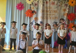 Dwoje dzieci występuje przed publicznością, trzymając mikrofony. Pozostali przedszkolacy stoją obok, uczestnicząc w programie artystycznym przygotowanym na zakończenie roku szkolnego. W tle znajdują się barwne dekoracje wykonane z papierowych kwiatów.
