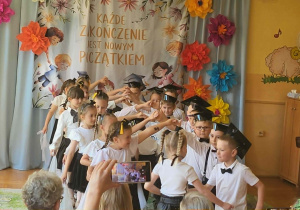 Starszaki podczas uroczystego występu kończącego rok przedszkolny. Dzieci stoją w rzędach przed dekoracją z napisem „Każde zakończenie jest nowym początkiem”, prezentując przygotowany program artystyczny.