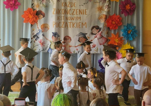 Przedszkolaki występują na scenie podczas uroczystości pożegnania przedszkola. Dzieci prezentują swoje umiejętności wokalne i recytatorskie, a zgromadzeni goście z uwagą oglądają przygotowany program.