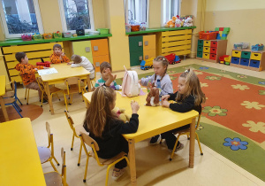 Dzieci siedzą przy stolikach, rozmawiają i bawią się podczas porannego dyżuru w sali przedszkolnej.