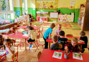 Dzieci siedzą przy stolikach i wykonują zadania w kartach pracy.