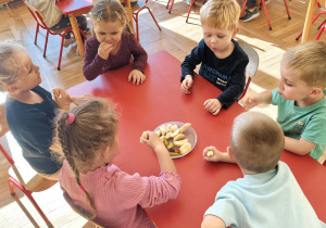 Przedszkolaki siedzą przy stolikach i z ciekawością degustują kawałki świeżych jabłek.
