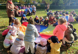 Dzieci stoją wokół kolorowej chusty animacyjnej i jednocześnie unoszą ją do góry. Jesienne liście podskakują na chuście, a przedszkolaki z radością obserwują efekty wspólnej zabawy.