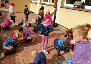Dzieci siedzą na chodniku i rysują kredą jesienne krajobrazy. Na nawierzchni powstają kolorowe rysunki inspirowane jesienną przyrodą.