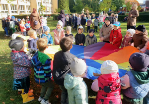 Dzieci stoją wokół kolorowej chusty animacyjnej i jednocześnie unoszą ją do góry.