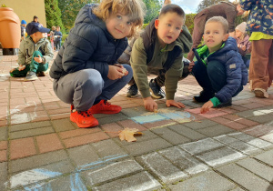 Dzieci siedzą na chodniku i rysują kredą jesienne krajobrazy. Na nawierzchni powstają kolorowe rysunki inspirowane jesienną przyrodą.