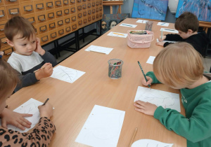 Dzieci uczestniczą w warsztatach plastycznych w bibliotece. Przy stolikach starannie malują i kolorują liście, tworząc barwne prace inspirowane jesienią.