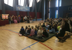 Dzieci z przedszkola siedzą na materacach w szkolnej sali gimnastycznej i oglądają przedstawienie teatralne „Królewska Akademia” w wykonaniu starszych uczniów.