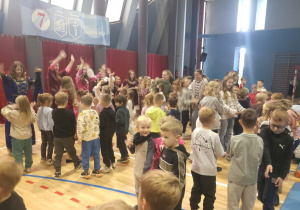 Uczestnicy spotkania stoją w kręgu i biorą udział w zabawie ruchowej, współpracując ze sobą i czerpiąc radość ze wspólnie spędzonego czasu.