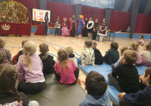 Uczniowie szkoły podstawowej prezentują sceny teatralne w pięknych strojach, a przedszkolaki uważnie oglądają występ w szkolnej sali gimnastycznej.