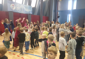 Przedszkolaki i starsi uczniowie biorą udział w tańcu integracyjnym, wykonując wspólne ruchy i gesty w rytm muzyki.