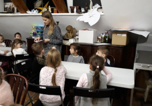 dzieci siedzą przy stolikach, pani tłumaczy dzieciom sposób wykonania pracy