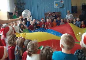 dzieci trzymają się za kolorowa chustę, pani przebrana w czerwony strój przebija w dłoniach balon z konfetii