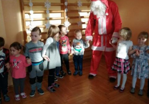 Mikołaj na tle drabinek gimnastycznych tańczy wspólnie z dziećmi trzymając je za ręce