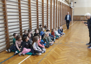 dzieci siedzą na materacach w szkolnej sali gimnastycznej, słuchają poleceń trenera, w tle widać dyrektora szkoły