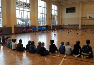 dzieci siedza tyłem do fotografa, w tle na podłodze leżą porozrzucane piłki