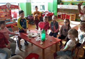 dzieci podczas eksperymentu siedzą wokół czerwonego stolika, na stole leży zielona butelka na której jest różowy balon