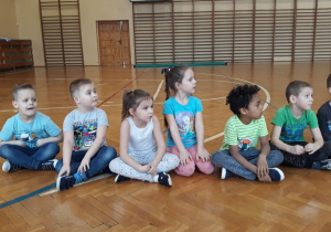 Sala gimnastyczna. Dzieci siedzą na podłodze, głowy zwrócone mają w prawą stronę, w tle widać drabinki