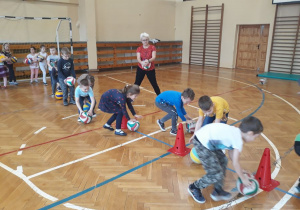 Sala gimnastyczna. Dzieci wykonują ćwiczenia z piłkami, toczą piłki wokół pachołków. Trener stoi z tyłu drużynyy