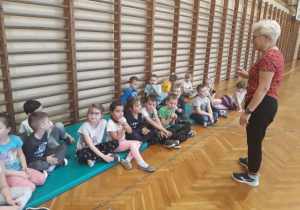 Szkolna sala gimnastyczna. Dzieci siedza na materacach, słuchają poleceń trenera, który stoi na przeciwko drabinek