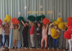 dzieci stoją z uniesionymi dłońmi w których trzymają kolorowe pompony