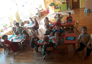 przedszkolaki siedzą przy stolikach, malują farbami koloru zielonego i niebieskiego szablon planety Ziemia
