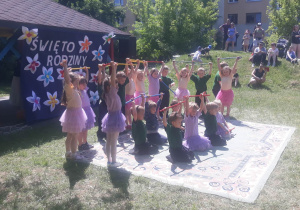 dzieci z grupy IV podczas występów, ubrane w fioletowo-różowe stroje trzymają w dłoniach laski gimnastyczne uniesione do góry