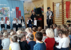 Pani Dyrektor przemawia do dzieci przez mikrofon, w tle stoją przedszkolaki ubrane w strój galowy