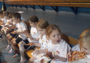 dzieci siedzą na trybunach, jedzą pizzę