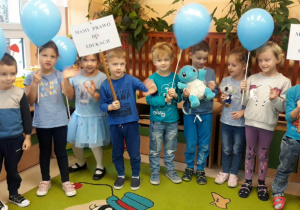 dzieci stoją na dywanie, trzymaja w dłoniach niebieskie baloniki oraz hasła