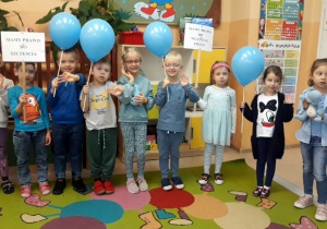 dzieci stoją na dywanie, trzymaja w dłoniach niebieskie baloniki oraz hasła