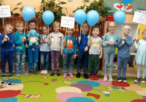 dzieci stoja na dywanie, trzymaja w dłoniach niebieskie baloniki oraz plakietki z napisami: Mamy Prawo do Edukacji, Mamy Prawo do Szczęścia, Mamy Prawo do Własnego Zdania
