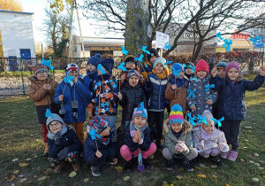 dzieci w ogrodzie przedszkolnym pozują do zdjęcia, w dłoniach trzymają niebieskie chorągiewki