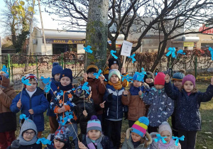 dzieci w ogrodzie przedszkolnym pozują do zdjęcia, w dłoniach trzymają niebieskie chorągiewki