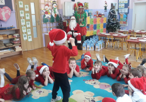 dzieci leżą na brzuchu, machają nogami chłopiec tańczy w środku, na głowie ma czerwoną czapkę Mikołaja