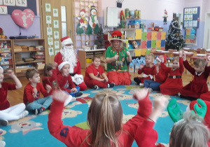 dzieci siedzą na dywanie z Mikołajem i elfem, śpiewają piosenkę