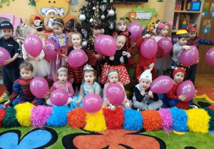 dzieci w strojach karnawałowych pozuja do zdjęcia, w dłoniach trzymaja różowe balony, na dywanie leżą kolorowe pompony